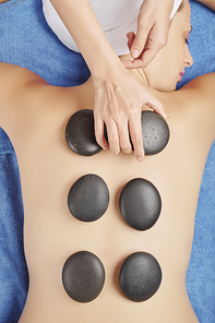 Hands of beautician putting hot round smooth heated rocks on back of young relaxed woman