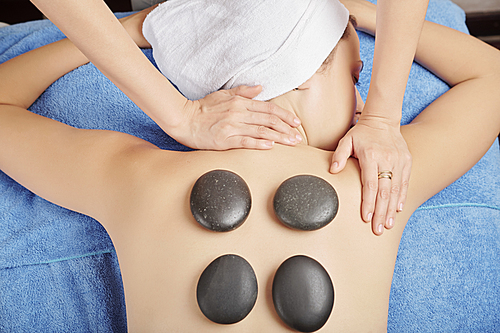 Masseur putting hot basalt stones on back of young woman and massaging her neck and shoulders