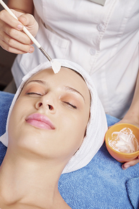 Close-up image of beautician applying white clay mask on face of pretty young woman