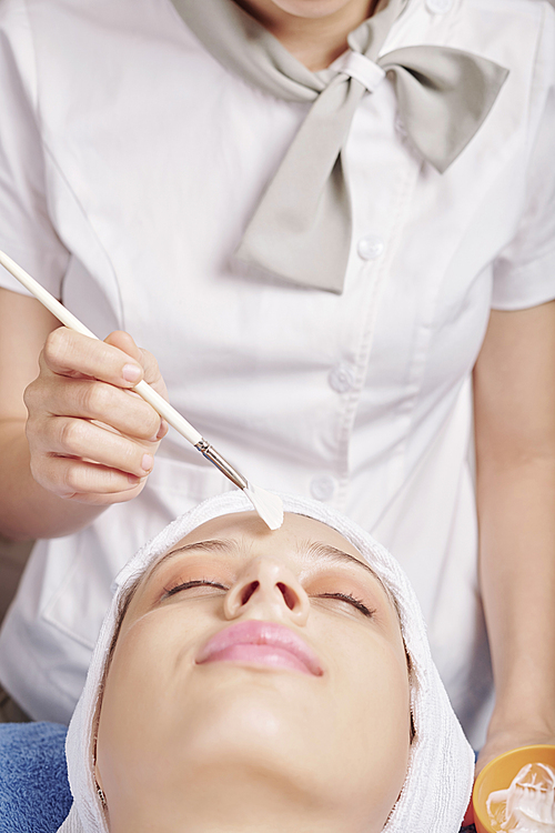 Beautician applying natural moisturizing face mask on forehead of relaxed female client