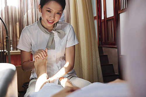 Young Vietnamese masseur giving professional relaxing feet massage to young femal client of spa salon