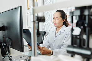 Asian mature woman sitting at the table in front of computer with folder and reading a medical card at office