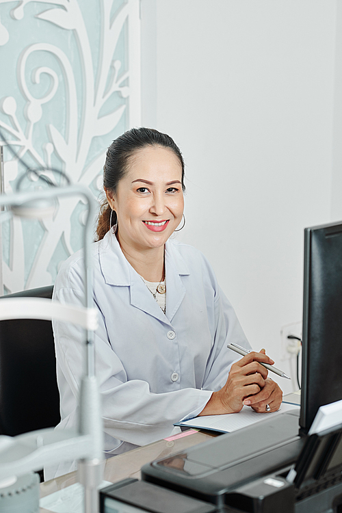 Portrait of Asian mature doctor in white coat smiling at camera while working at the table with documents at office