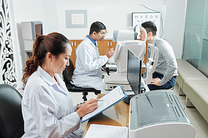 Asian nurse in white coat sitting at the table and filling the medical card while doctor examining the patient in the background at hospital