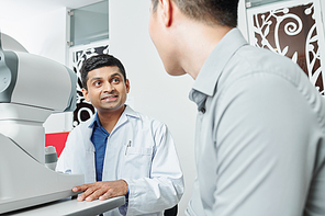 Indian doctor in white coat sitting at the table in front of medical equipment and talking to the patient during his visit at office