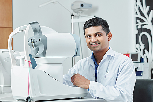 Portrait of Indian mature doctor in white coat smiling at camera while sitting at the table in front of modern medical equipment at hospital