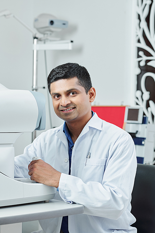 Portrait of Indian male doctor in white coat sitting at his workplace and smiling at camera at office
