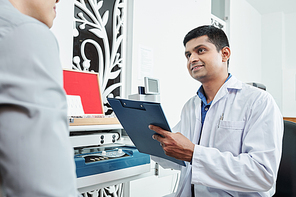 Indian doctor in white coat holding medical card and talking to the patient during his visit to the hospital