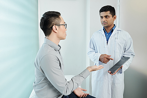 Indian doctor in white coat pointing at digital tablet in his hand and talking to the patient at office