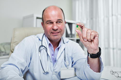 Smiling mature general practitioner showing best pain piller pill in his hand, selective focus