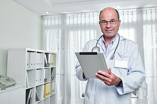 Portrait of smiling mature general practitioner in glasses holding tablet computer and looking a camera