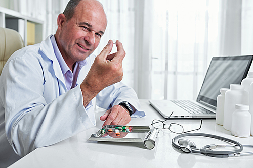 Smiling mature general practitioner looking at tablet of effective painkiller in his hand