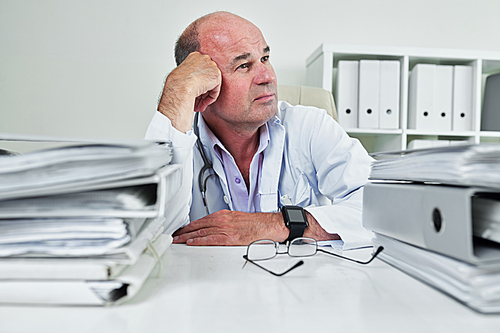 Mature general practitioner sitting at his table piled with binders and documents and daydreaming
