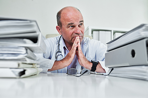 Serious experienced general practitioner holding hands together in praying gesture and hoping for the better