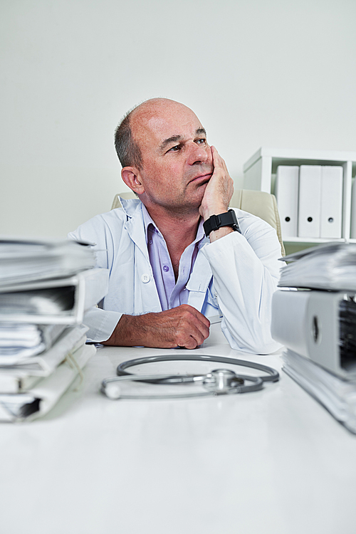 Portrait on pensive mature general practitioner sitting at his office table piled with binder and folders of medical documents