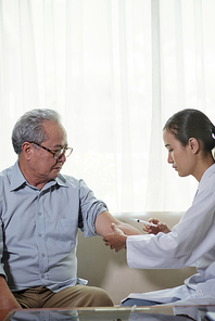 Young nurse in white coat sitting on sofa and make an injection in hand to her senior patient at home