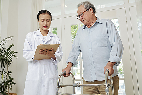 Asian young doctor in white coat standing and making notes in document with senior patient standing with walker and listening to her recommendation