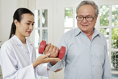 Senior Asian patient in eyeglasses exercising with dumbbells with Asian young nurse in white coat helping to him he recovering after operation