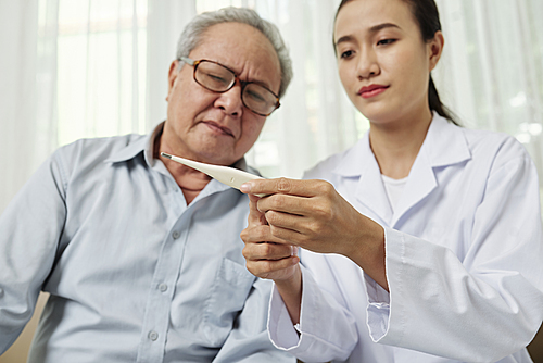 Asian young nurse in white coat sitting together with her senior patient holding thermometer and showing him the temperature of his body