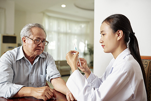 Asian female doctor sitting at the table and is going to make a vaccination to the senior man during her home visit