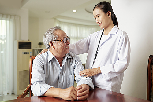 Asian young nurse standing in white coat and care about senior man while he sitting at the table and talking to her at home