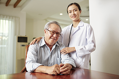 Portrait of young nurse in white coat standing near the senior man in eyeglasses who sits at the table and they smiling at camera
