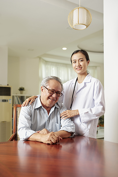 Portrait of happy senior man sitting at the table with young female doctor standing near by him they smiling at camera at doctor's office