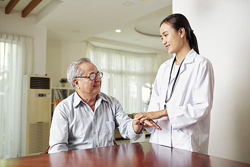 Asian female doctor in white coat holding hand of her senior patient and smiling while he sitting at the table and talking to her at hospital