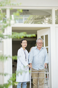 Portrait of senior man standing with walker and  together with young nurse in white coat they standing near the door