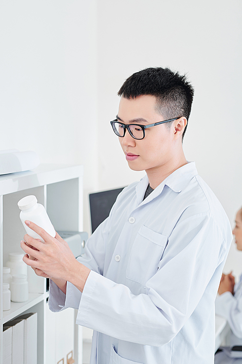 Young Asian pharmacist in white labcoat taking container of pills and tablets from the shelf and reading information on it