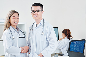 Portrait of smiling young Vietnamese general practitioners in white labcoats, their colleague working on computer in background