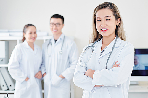 Pretty confident young smiling Vietnamese general practitioner with stethoscope standing in front of her colleagues