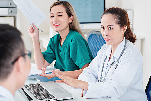 Serious middle-aged Vietnamese cardiologist talking to surgeon and her assistant at office table