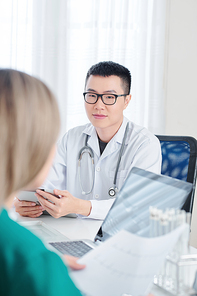 Young Vietnamese doctor in glasses sitting at table with digital tablet in hands and talking to physician