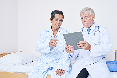 Doctor with tablet computer sitting on bed of senior patient and discussing with him therapy results