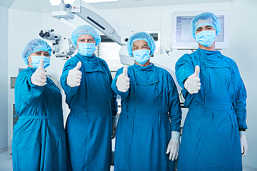 Team of surgeons in scrubs and face masks showing thumbs-up after suvcccessful surgery