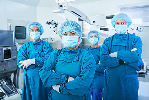 Smiling Vietnamese female chief surgeon standing in front of her team, folding arms and 