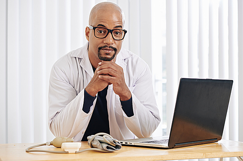 Portrait of mature doctor in white labcoat sitting at desk with opened laptop and 