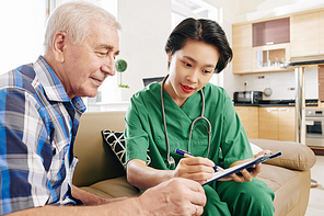 Young Vietnamese nurse vising senior patient at home and helping his with understanding of results of medical test