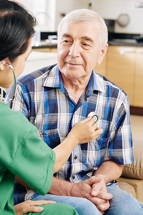 Smiling elderly man looking at hospital worker in green scrubs listening to his heartbeat when visiting him at home