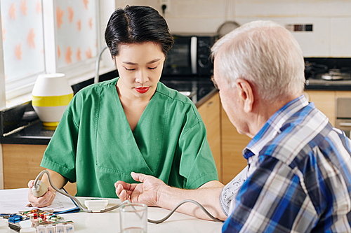 Serious young Asian medical nurse using tonometer to measure blood pressure of senior patient when visiting him at home