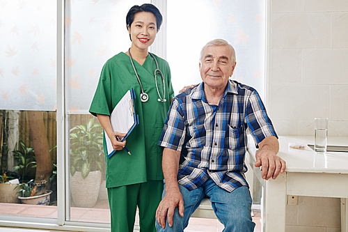 Portrait of smiling senior man sitting next to his medical nurse visiting him at home for weekly check-up