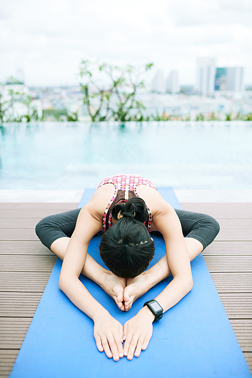 Young flexible woman exercising on exercise mat with swimming pool in the background in summer day outdoors