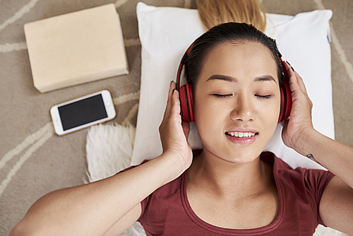 Face of pretty young Asian woman closing eyes and listening to music in headphones, view from above