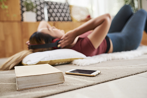Young woman resting on the floor and listening to music in headphones, her smartphone and thick book on foreground