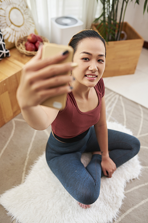 Smiling beautiful young Vietnamese woman sitting on the floor in her room and taking selfie