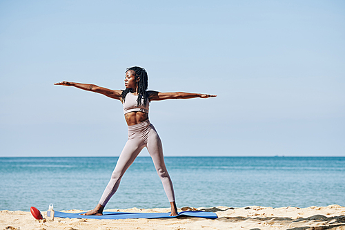 Slender beautiful young Black woman standing in warrior pose on yoga mat on the beach