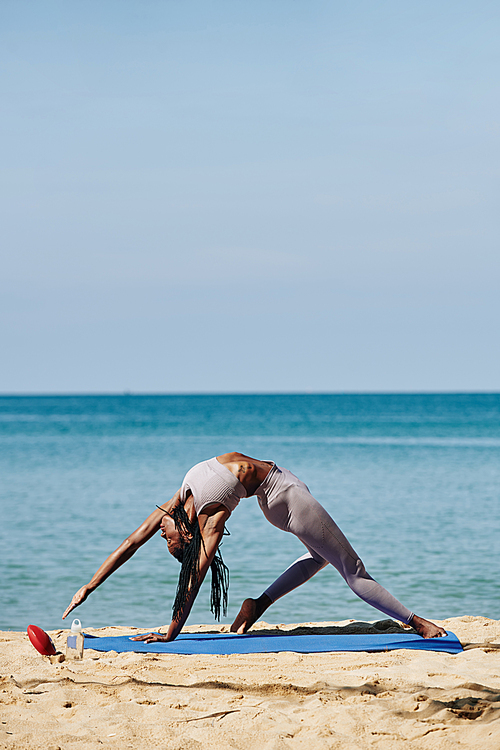 Flexible young Black woman practicing yoga and stretching outdoors by the ocean
