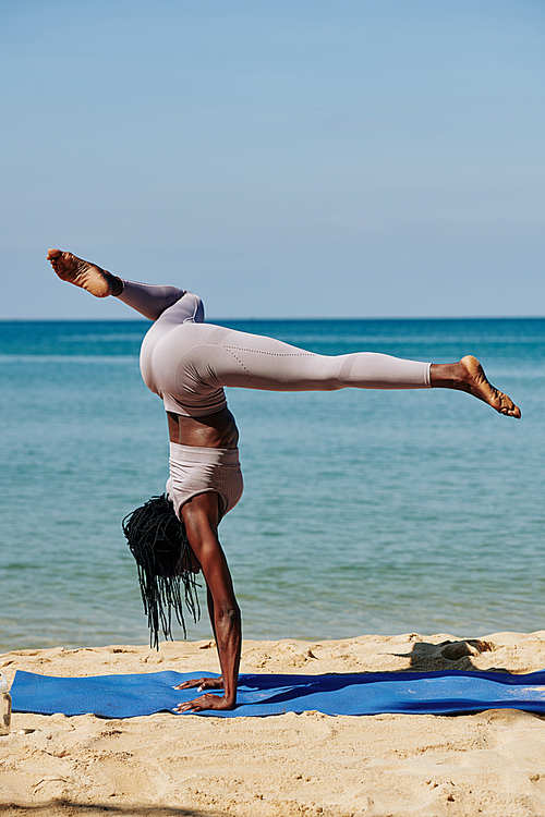 Slim young Black woman working out by the ocean and practicing handstand