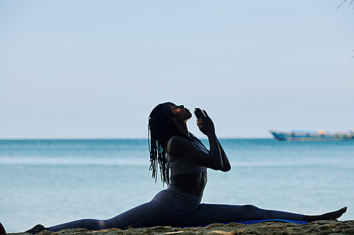 Young woman doing splits and keeping hands in namaste gesture, beautiful sea in background